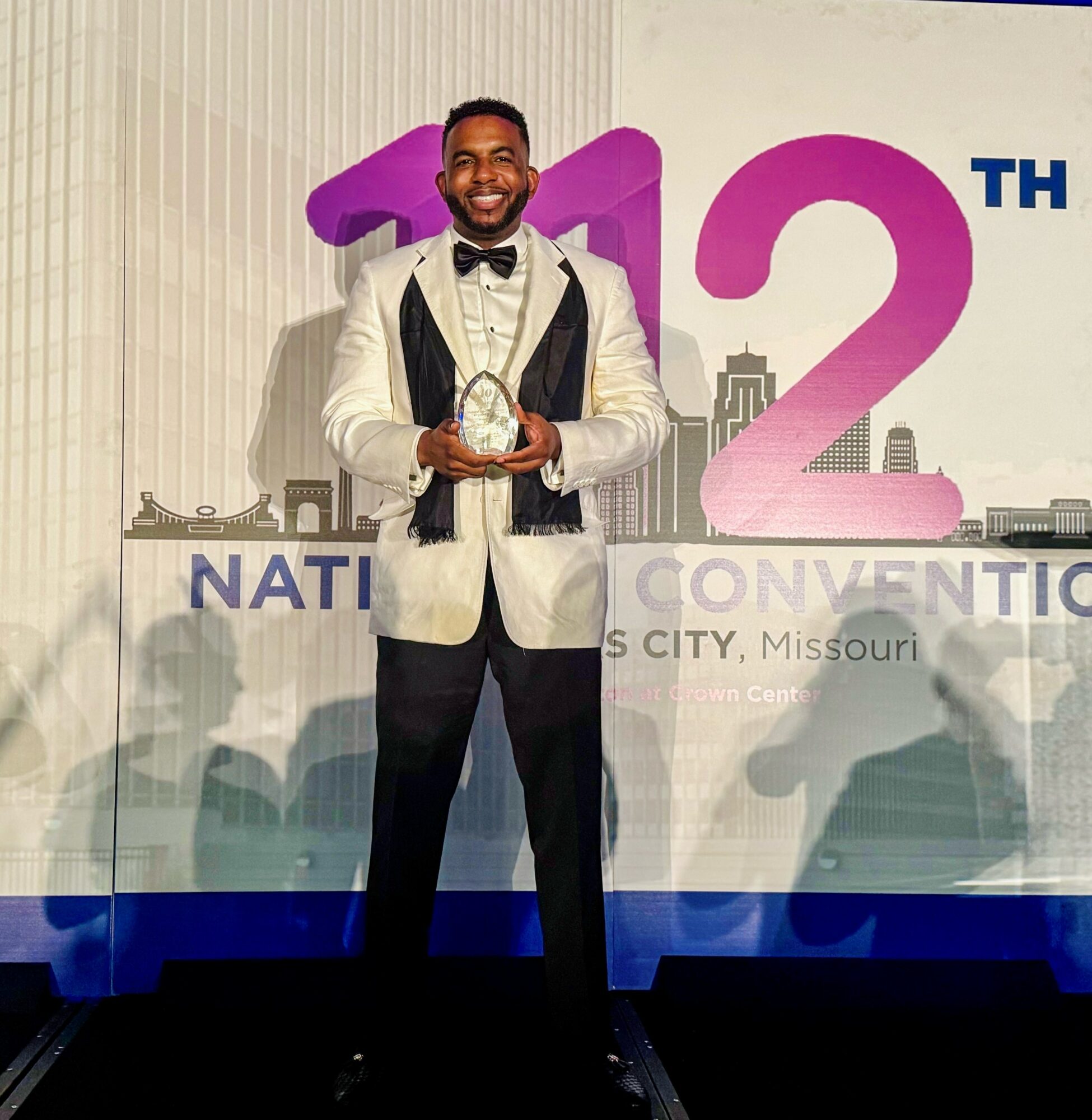 Man in tuxedo holding an award, standing in front of a large event backdrop with text and cityscape.