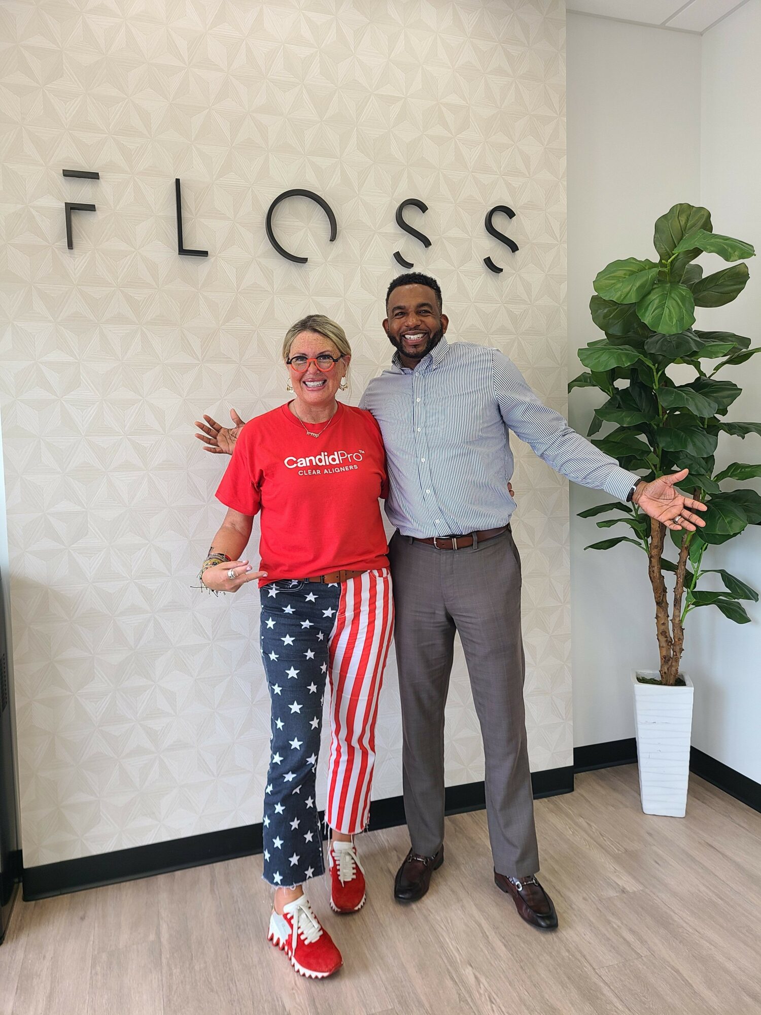 Two people standing in front of a wall with the word 'FLOSS'. One wears a red shirt and star-spangled pants, the other in a light blue shirt and gray pants.