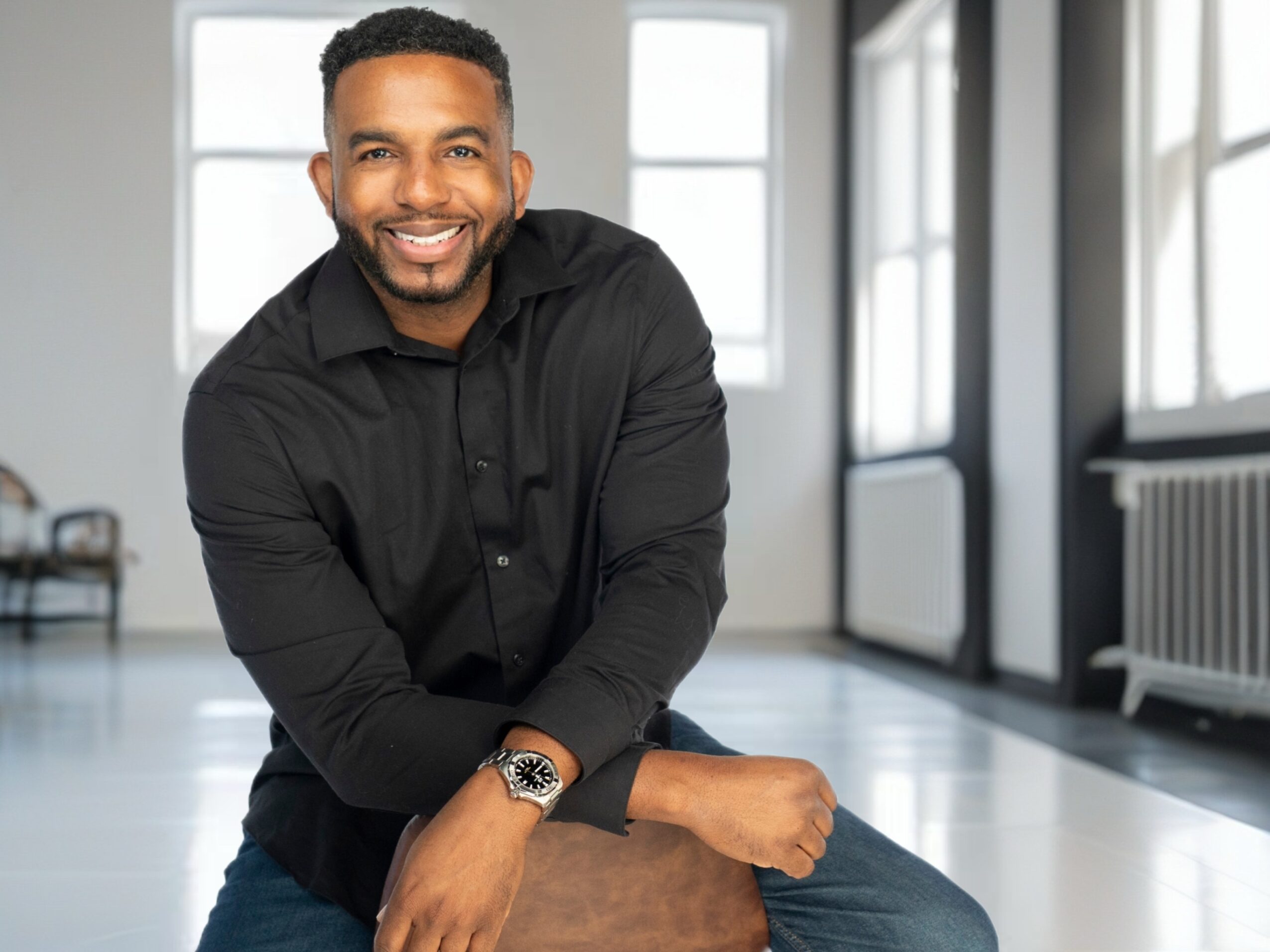 Smiling man with short hair and beard, wearing black shirt, sitting indoors near windows.