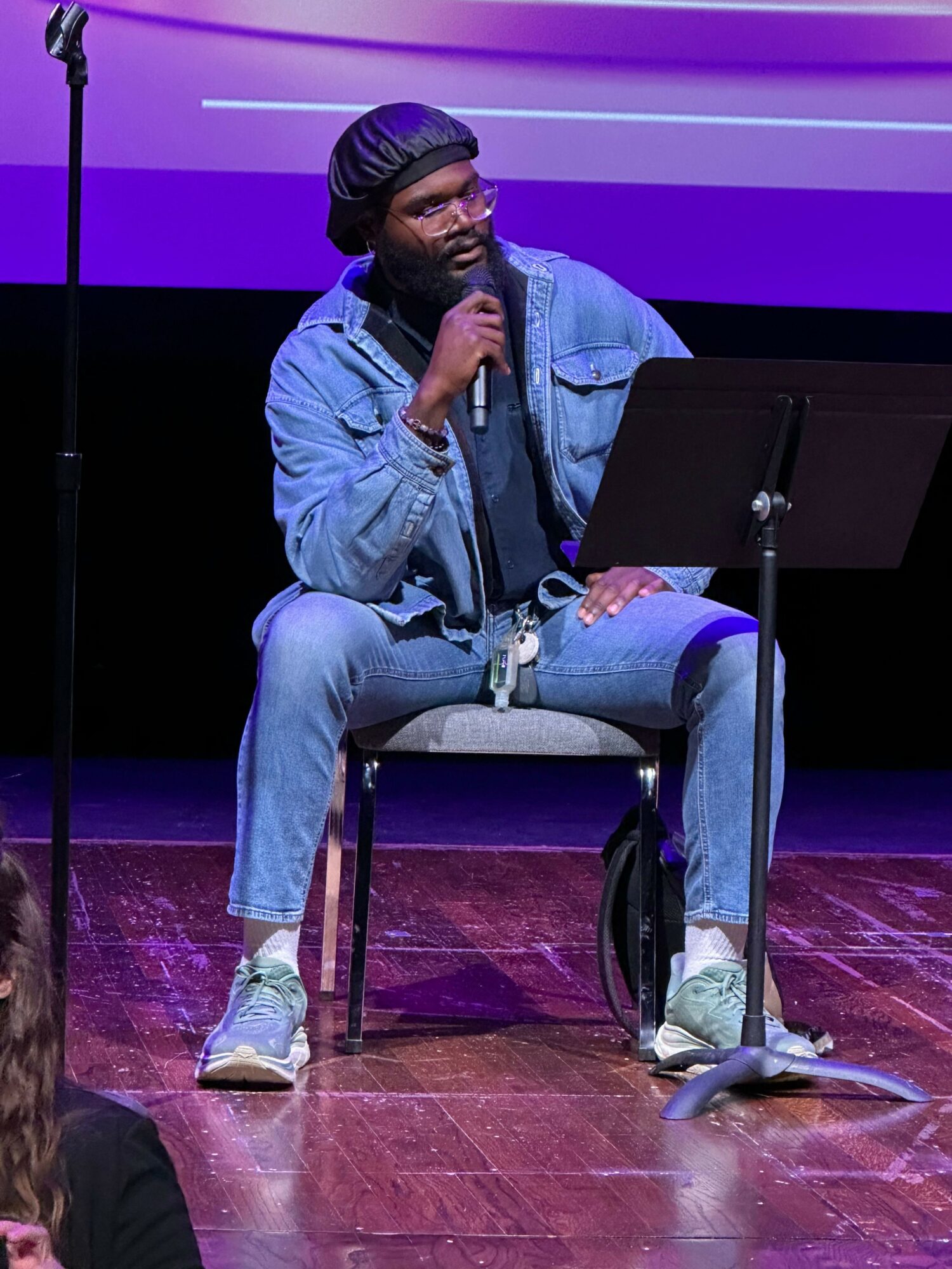 Person sitting on chair, holding microphone, wearing denim jacket, jeans, sneakers, and a beanie, on stage with purple background.
