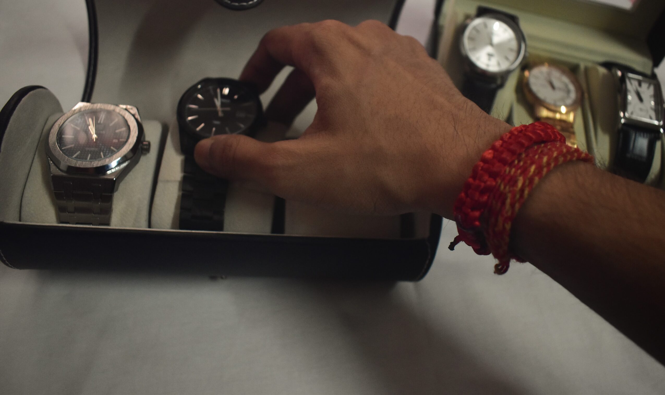 Person's hand reaching for a watch on a tray with multiple watches, wearing a red bracelet.