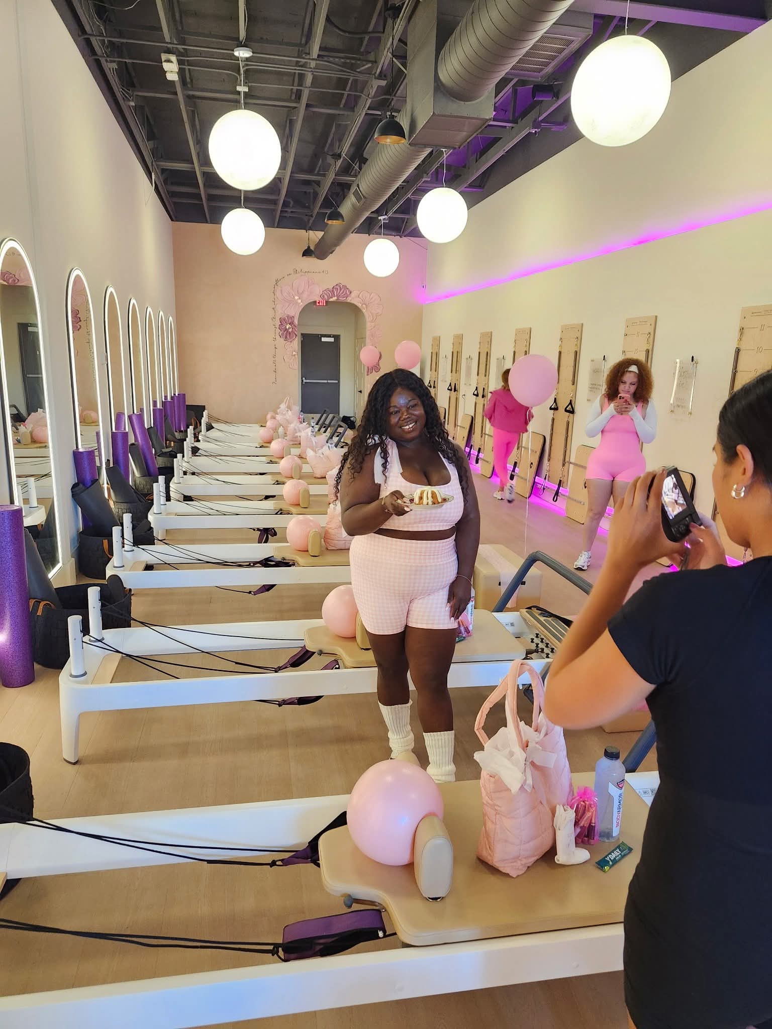 Indoor space with exercise equipment, pink balloons, and people, including a woman taking a photo, in a modern studio.