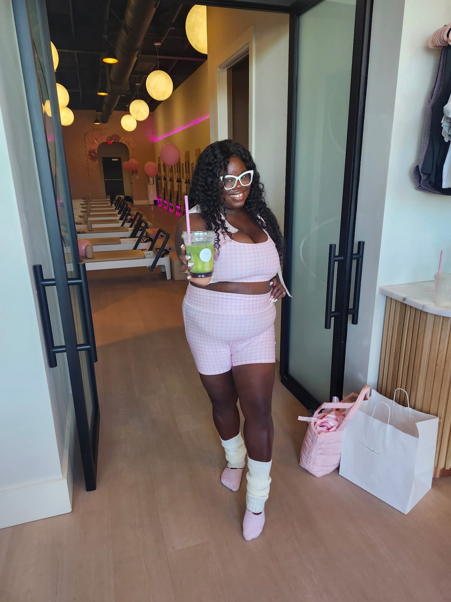 Woman with curly hair and sunglasses holding a drink, standing in doorway inside a modern cafe or restaurant.