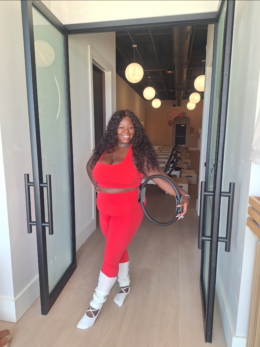 Woman in red jumpsuit and white boots standing in doorway, holding a black circular object, smiling, with hanging lights inside.