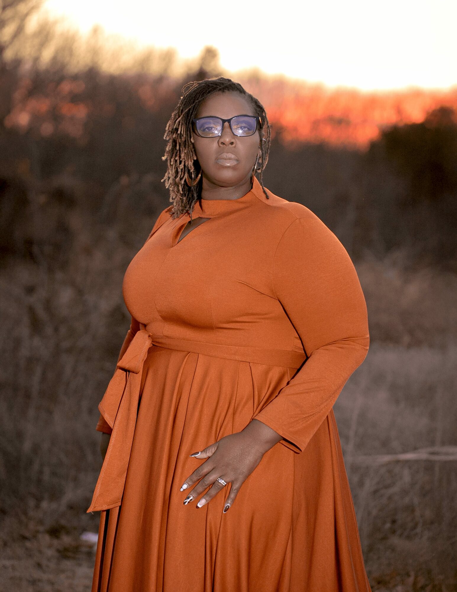 Woman in orange dress and sunglasses outdoors during sunset, standing with hand on hip, landscape background.