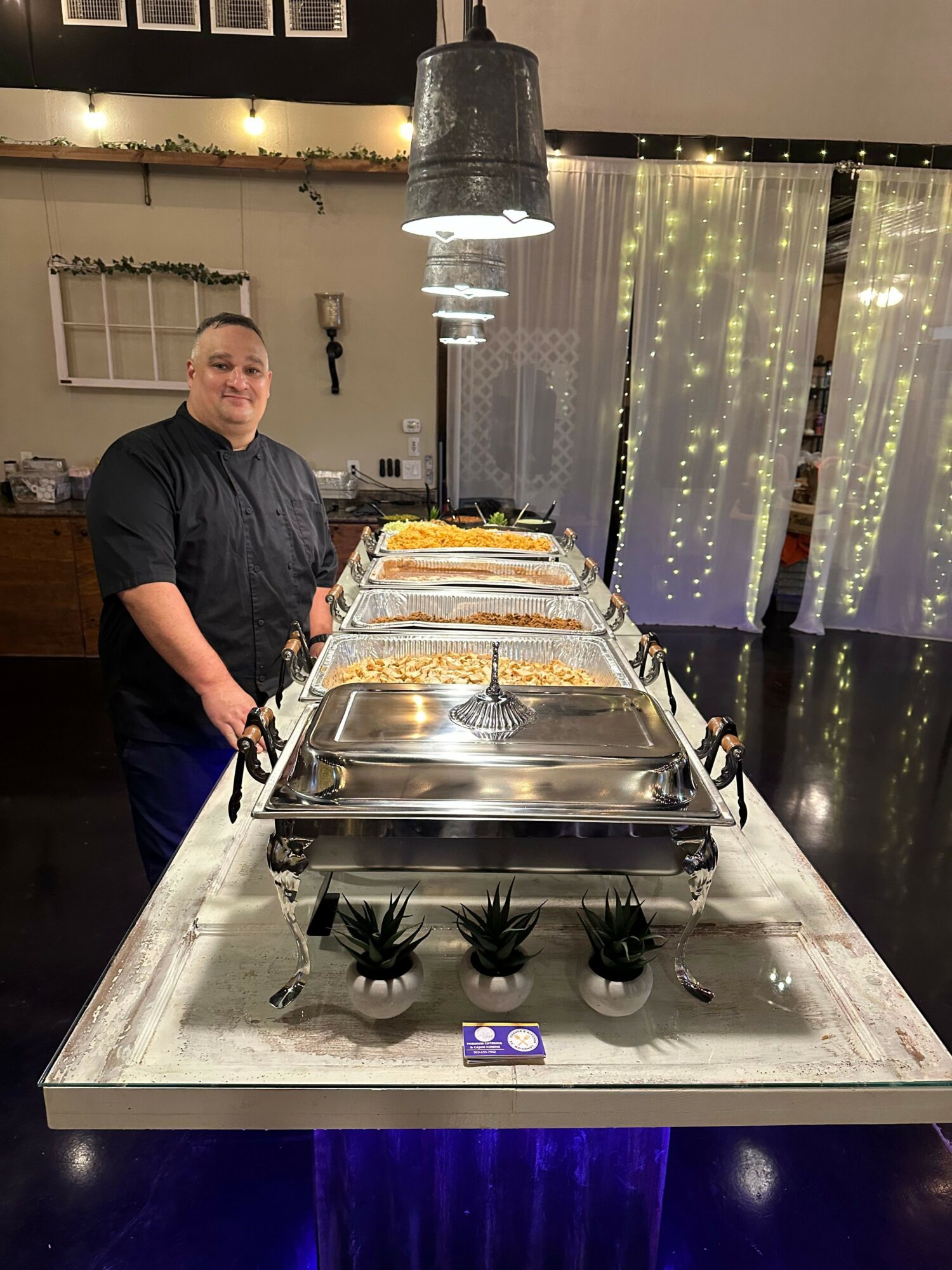 Chef standing behind a buffet table with covered dishes, small plants, and a digital timer, in a decorated dining area.