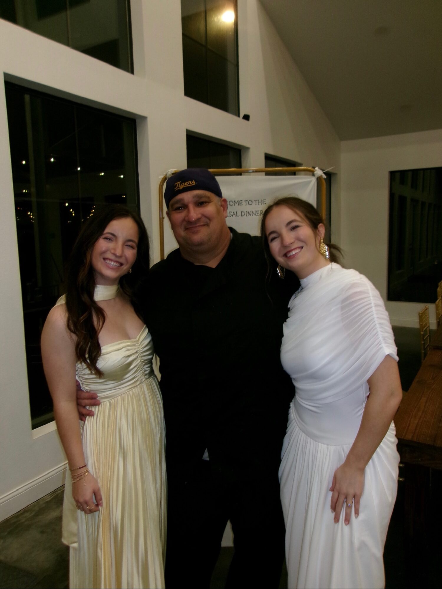 Three people standing indoors, two women in white dresses and a man in black, smiling at the camera.
