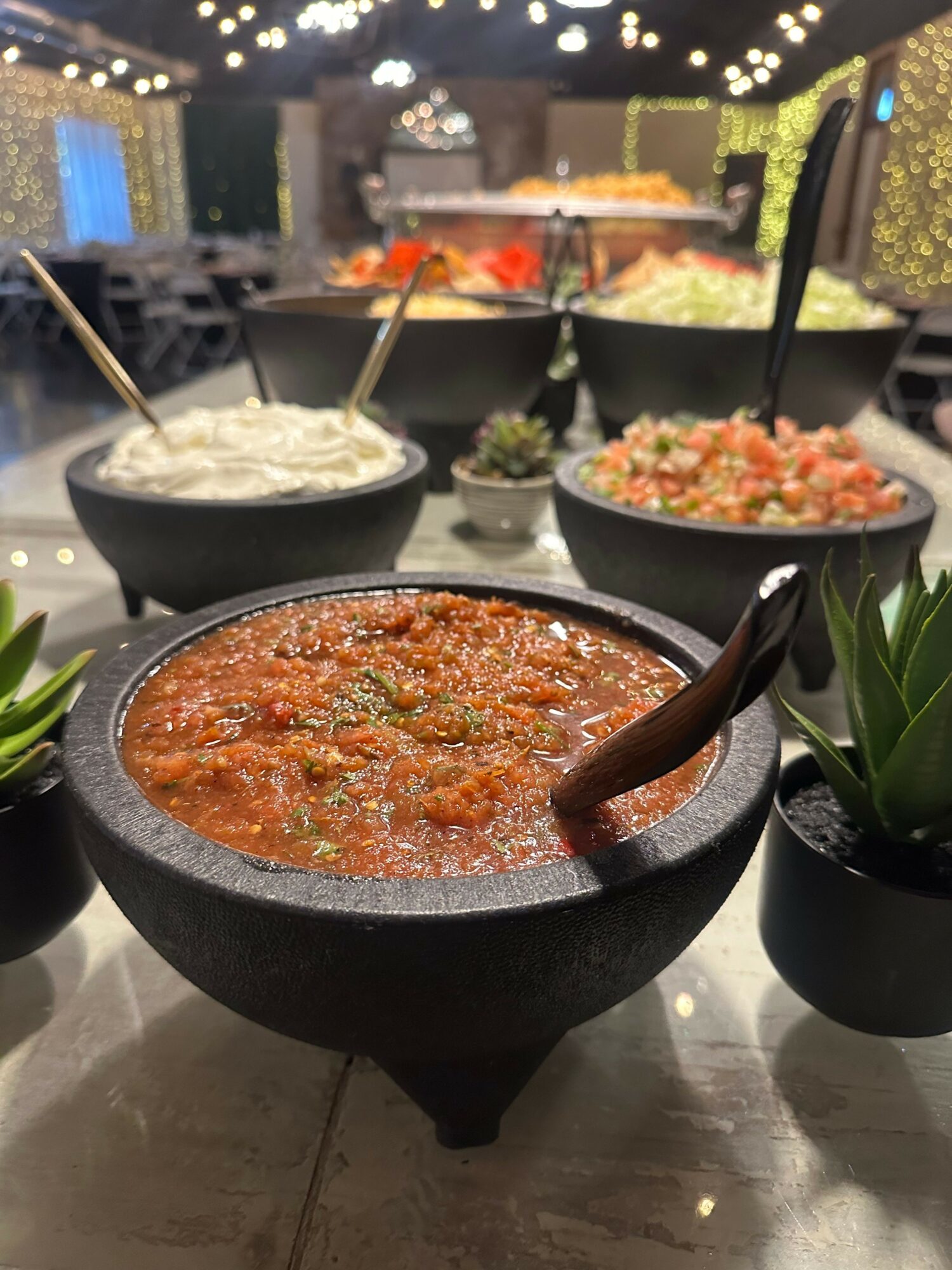 Bowls of various dishes on a table with decorative plants, blurred background of a restaurant or event space.