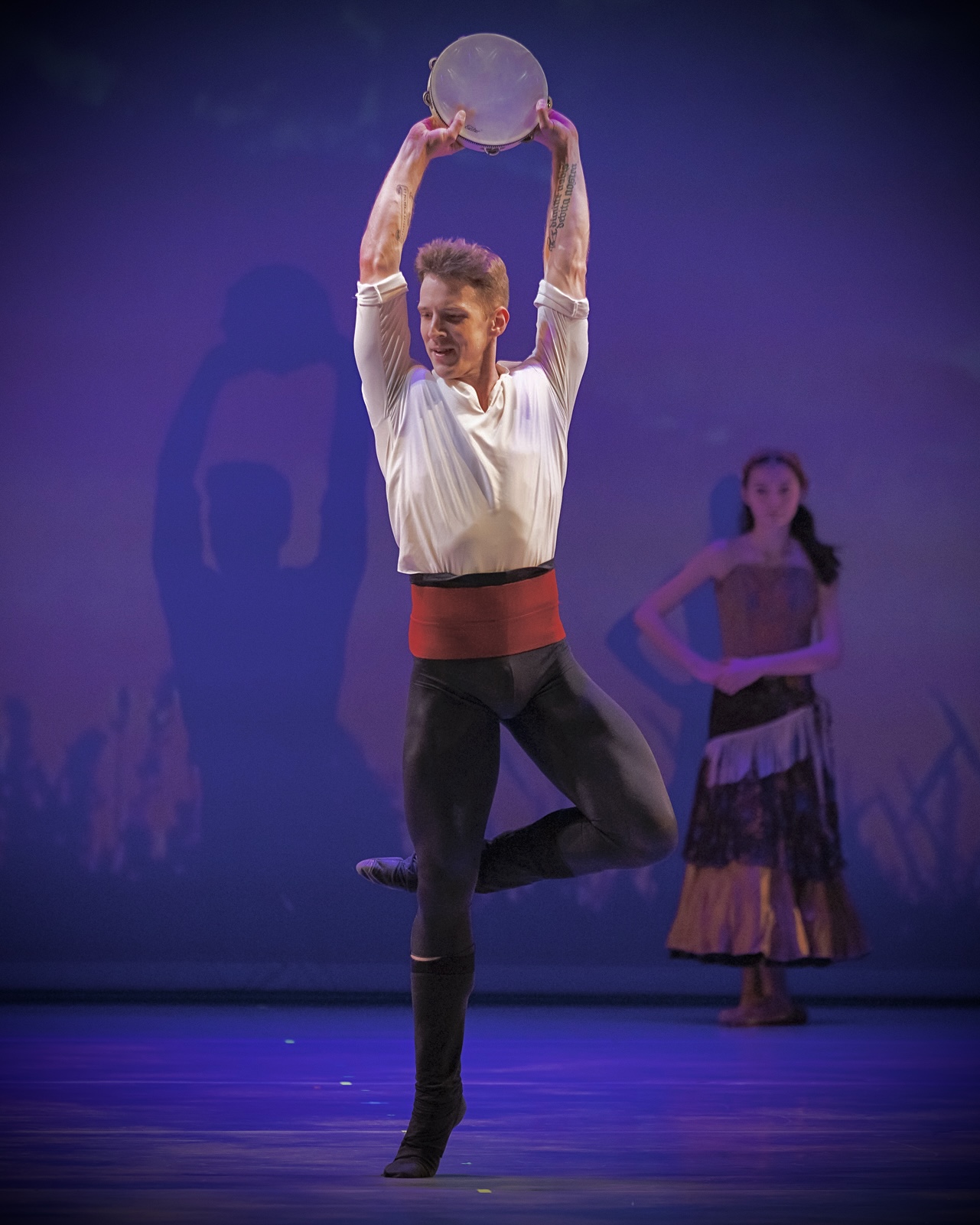 Performer balances on one leg while holding a circular object above head, with another performer in background.