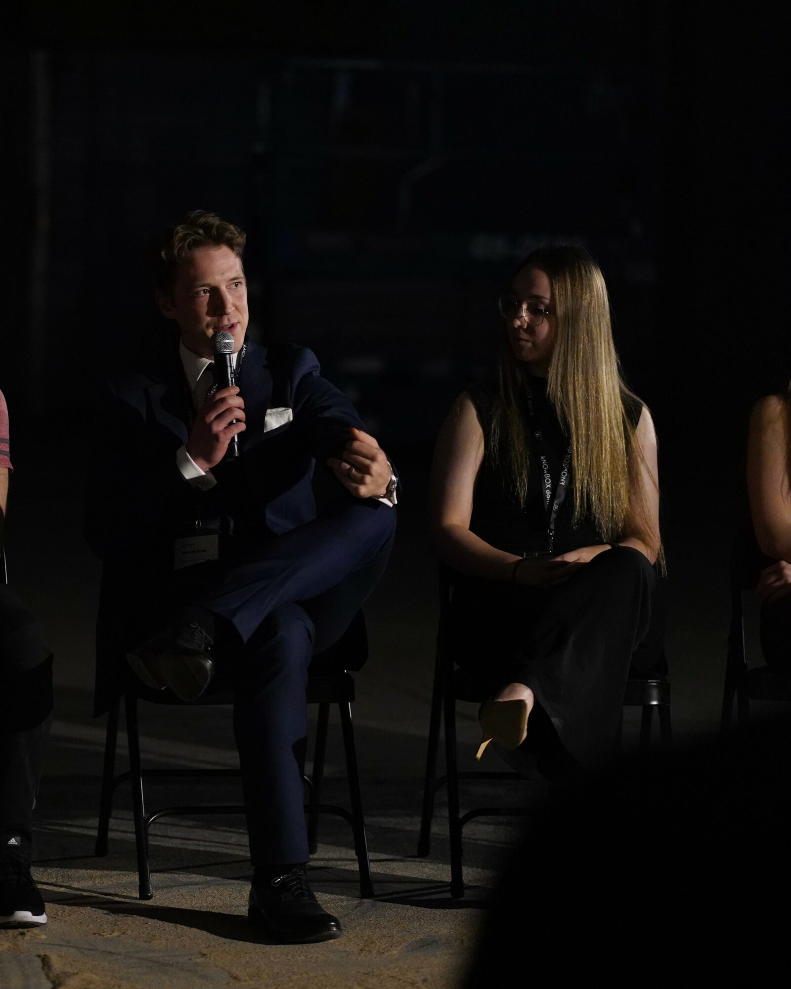 Man speaking into microphone and woman sitting on stage, both in dark clothing, in a dimly lit setting.