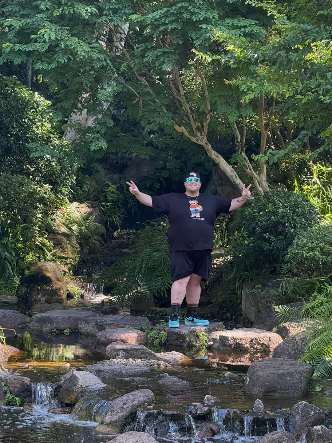 Person standing on rocks in a stream with arms outstretched, surrounded by lush green trees and foliage.