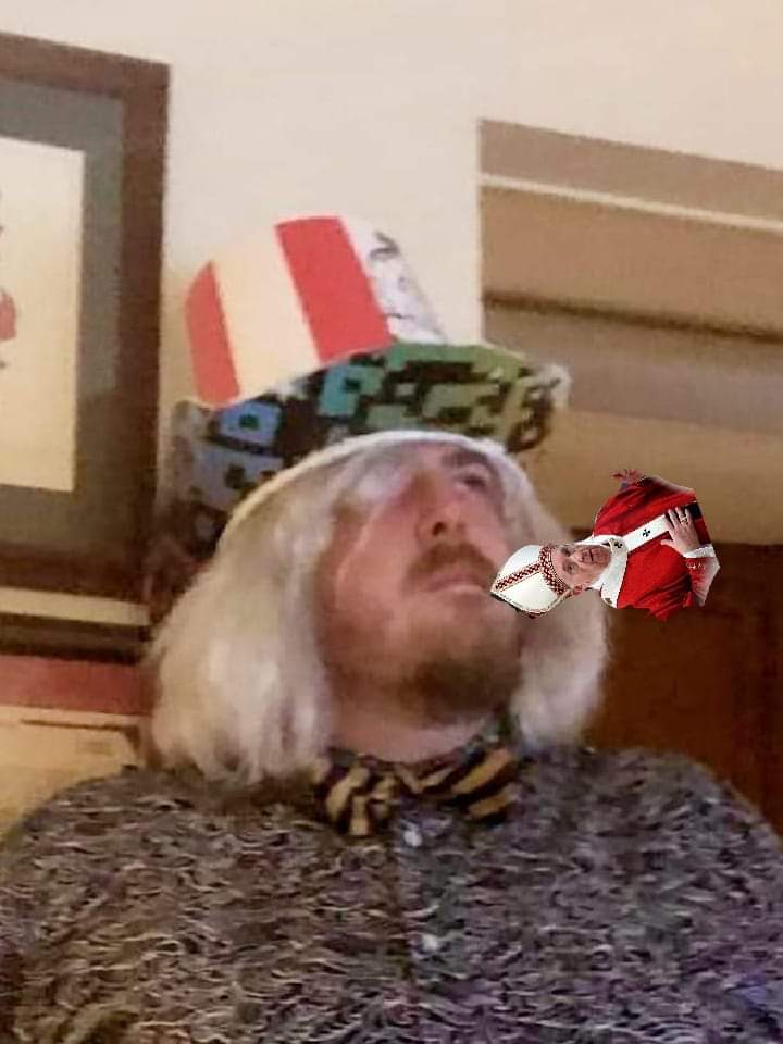 Person with long blonde hair wearing a patterned shirt and bow tie, holding a toy gun to their head, in a room.