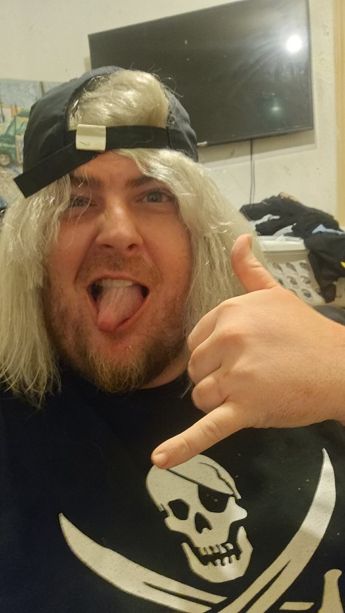 Person with long blonde hair wearing a black cap backwards, making a hand gesture, smiling with tongue out.