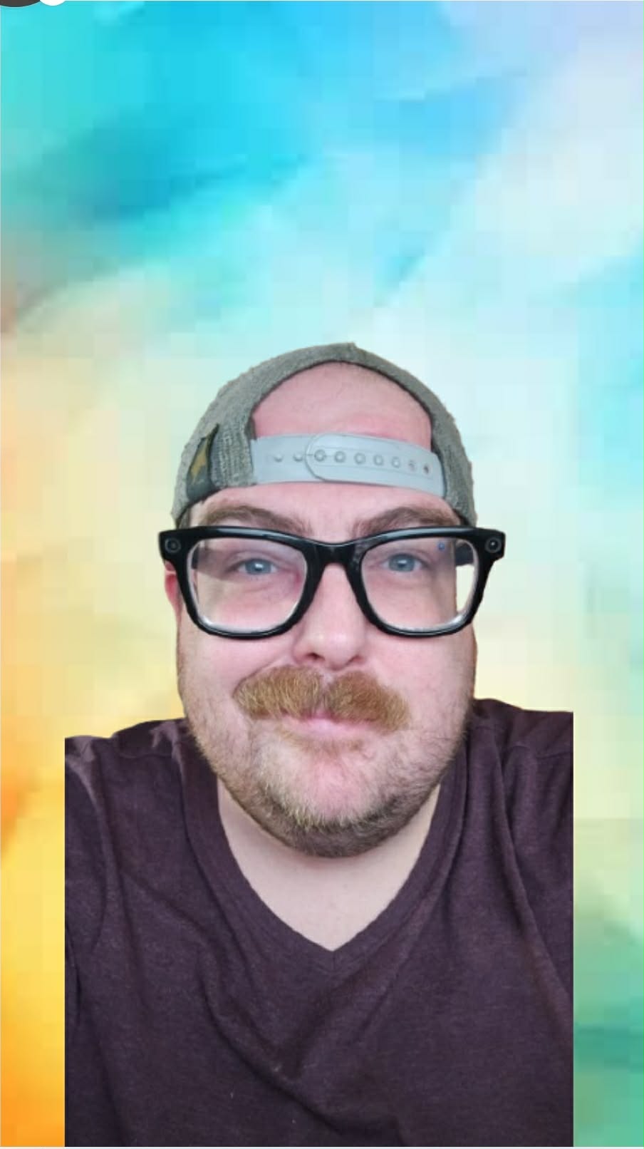 Man with glasses, cap backwards, mustache, smiling, wearing a dark shirt, colorful background.