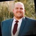 Man in a suit smiling outdoors with trees in background.
