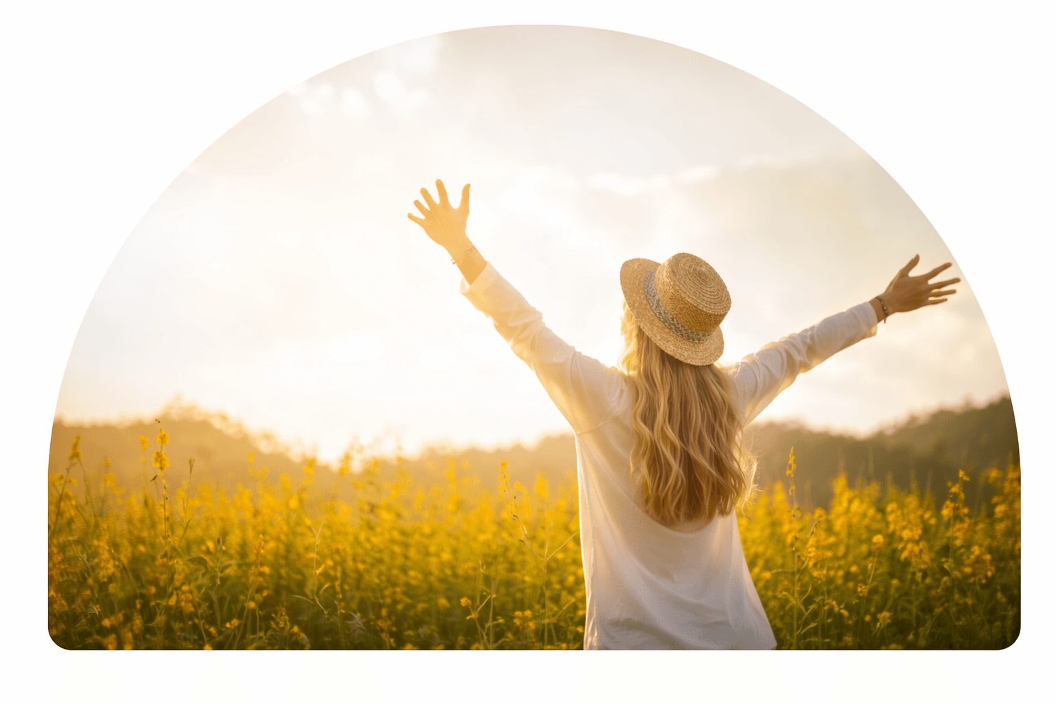 Person with long hair and a hat standing in a field with arms raised, facing away, during sunset.