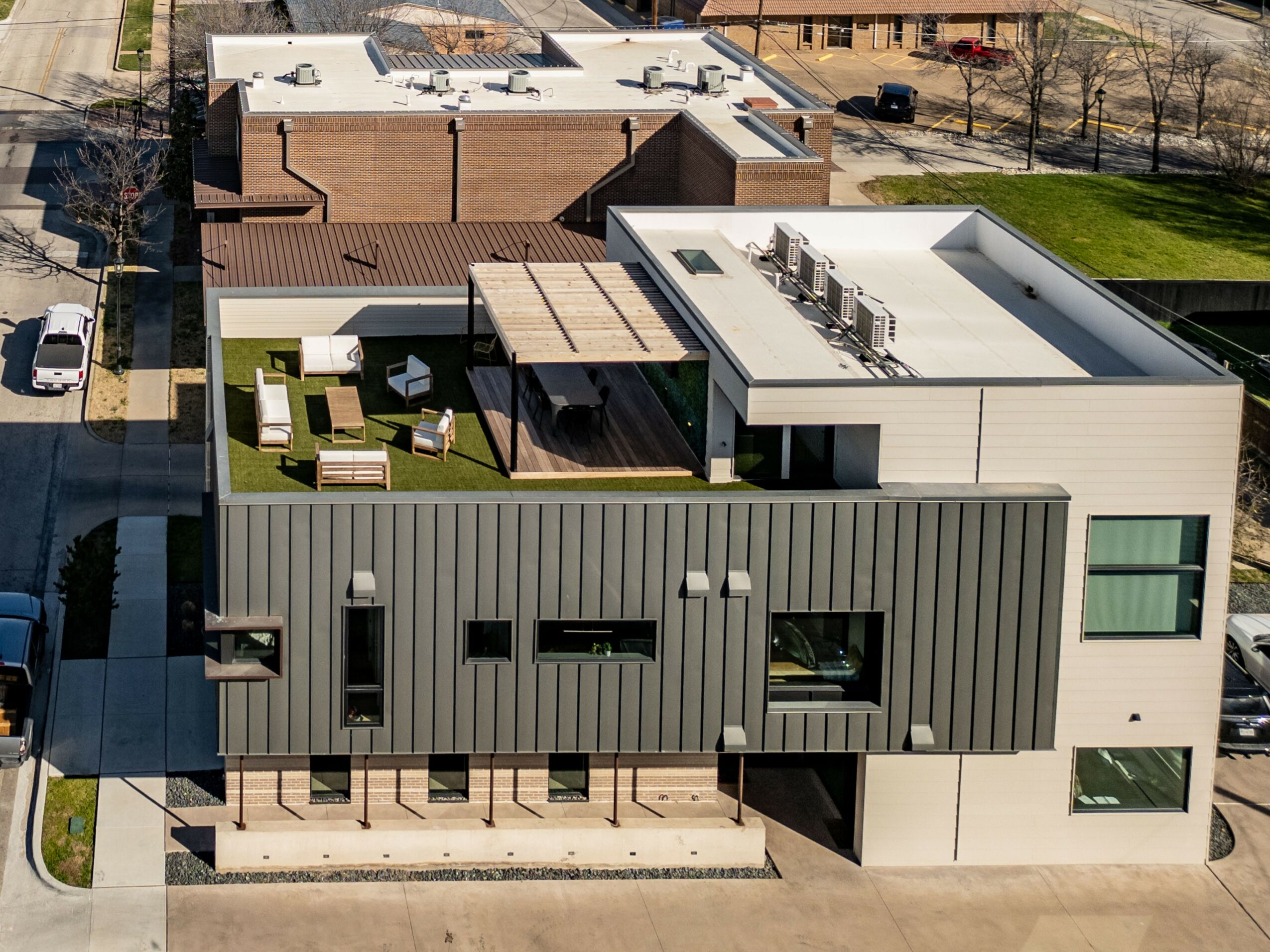 Modern building with outdoor seating area, rooftop equipment, and multiple windows, surrounded by trees and other buildings.