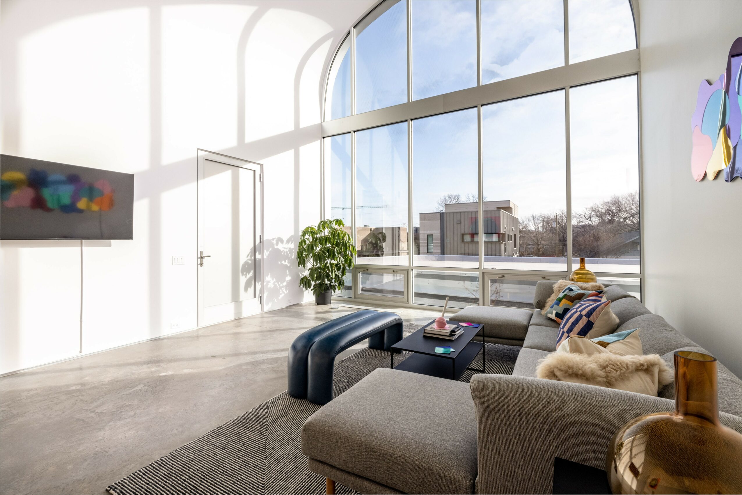 Modern living room with large windows, gray sofa, colorful pillows, black coffee table, and a potted plant.