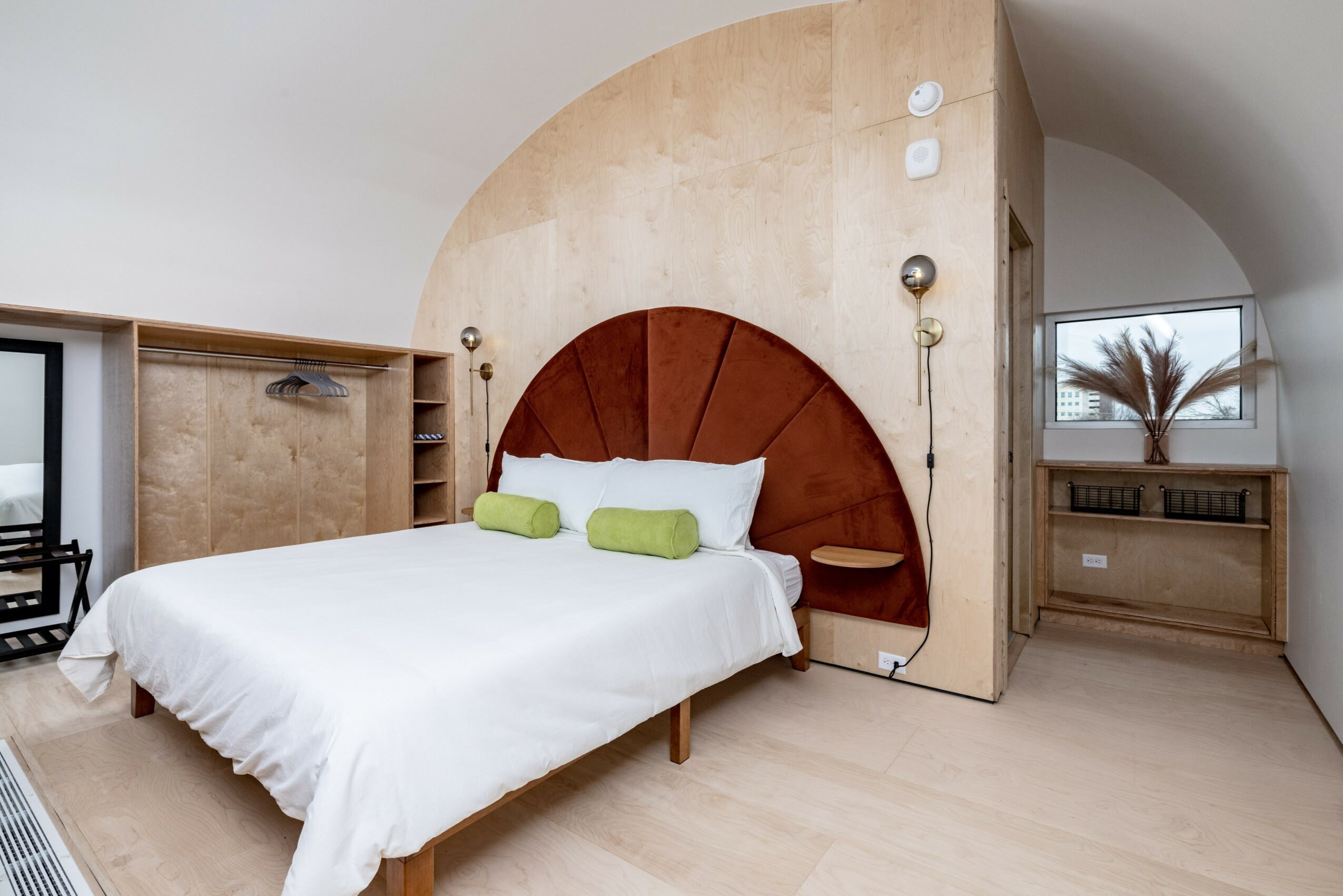 Bedroom with a bed, green pillows, wooden headboard, curved wall, and a window with a plant outside.