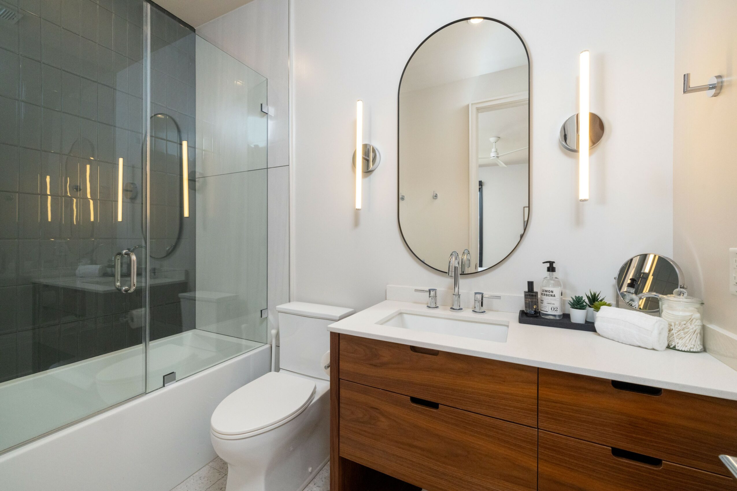 Bathroom with a glass shower enclosure, toilet, oval mirror, wooden vanity, and wall-mounted lights.