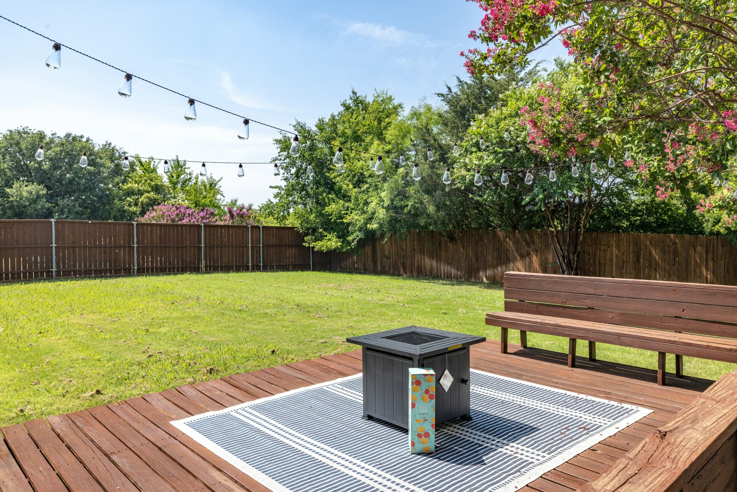 Backyard with wooden deck, grassy lawn, trees, and string lights overhead.