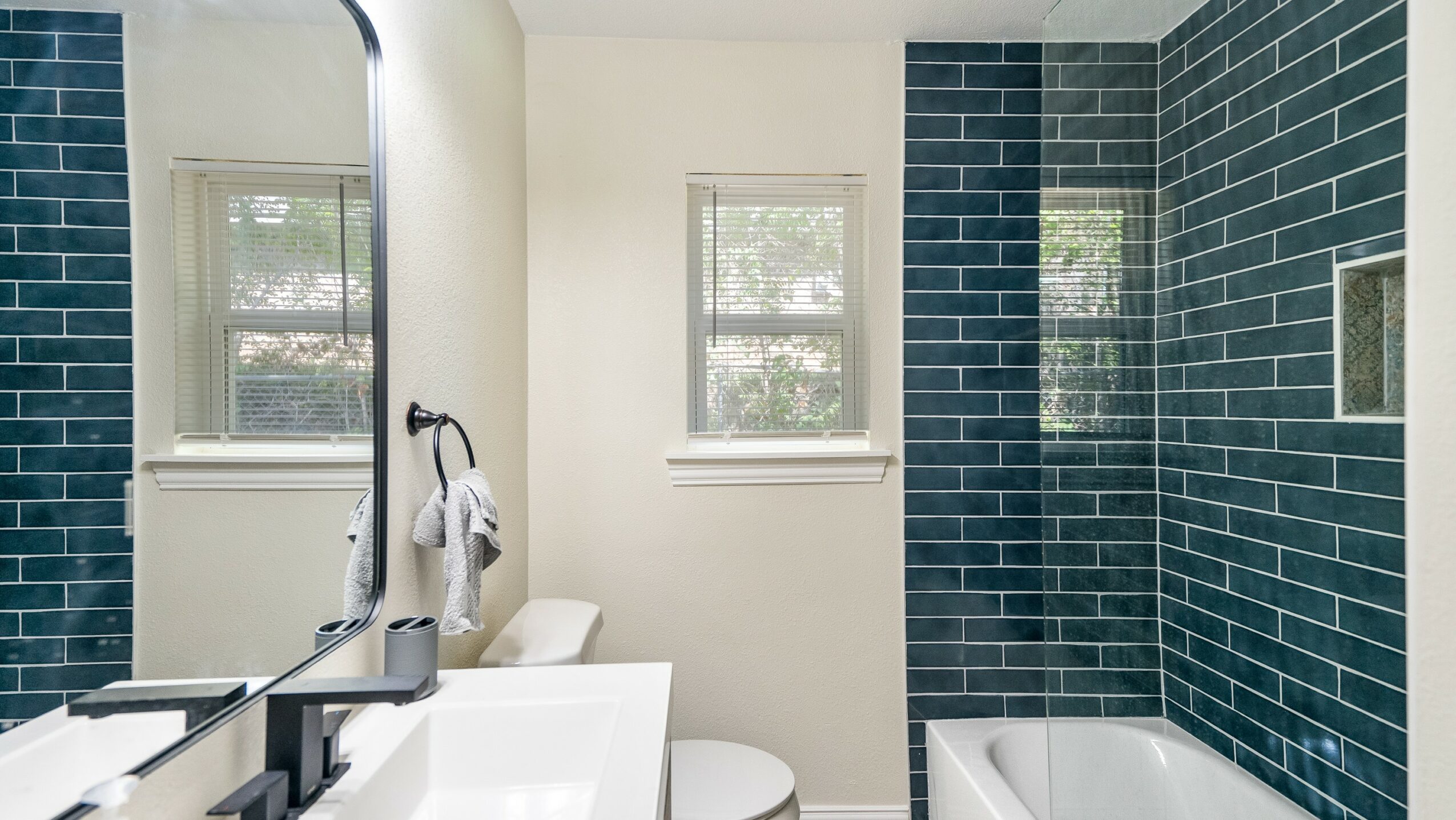 Bathroom with white walls, two windows, a mirror, and a shower area with dark blue tiles.