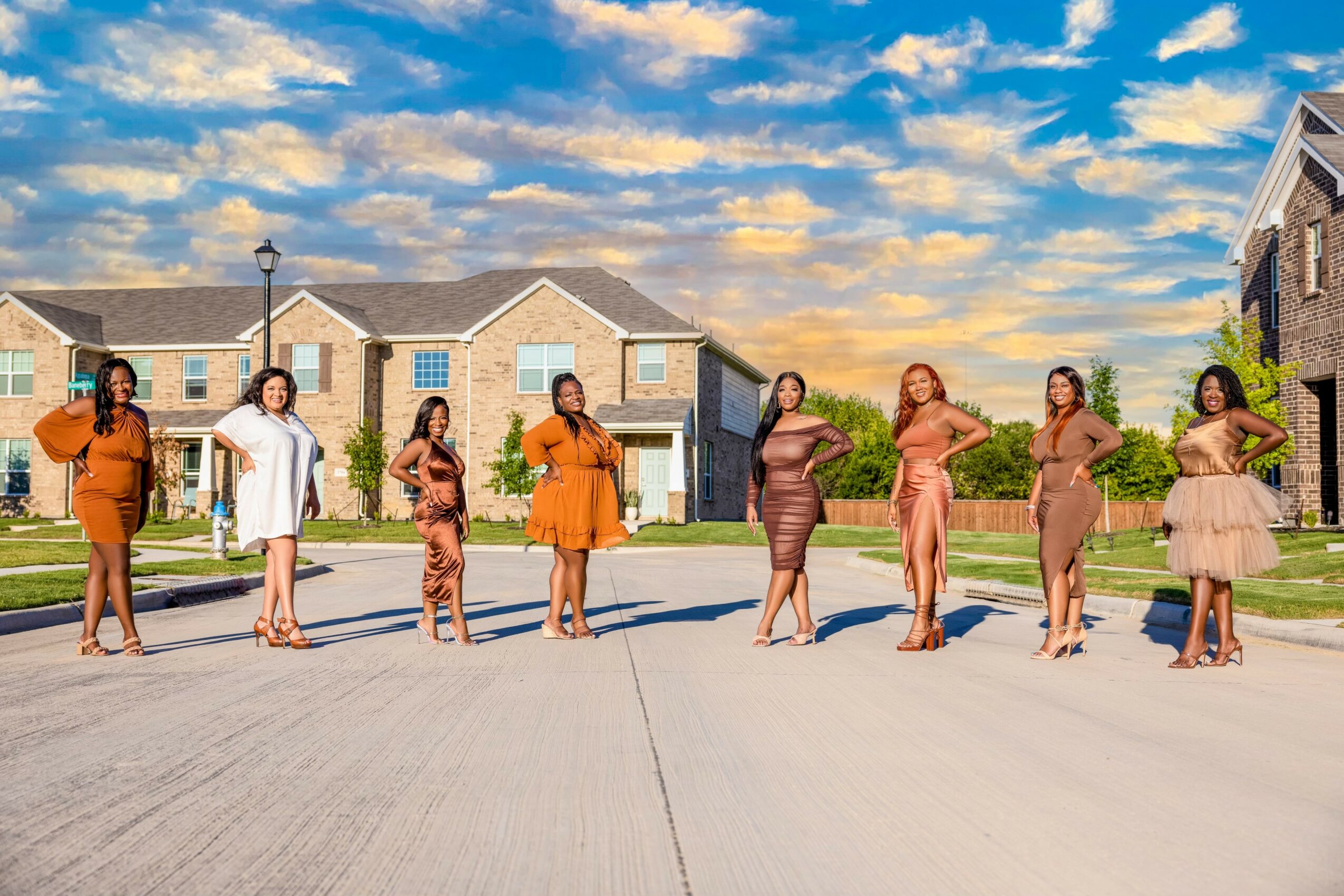 Eight women stand outdoors in a row on a street, with residential buildings and a partly cloudy sky in background.