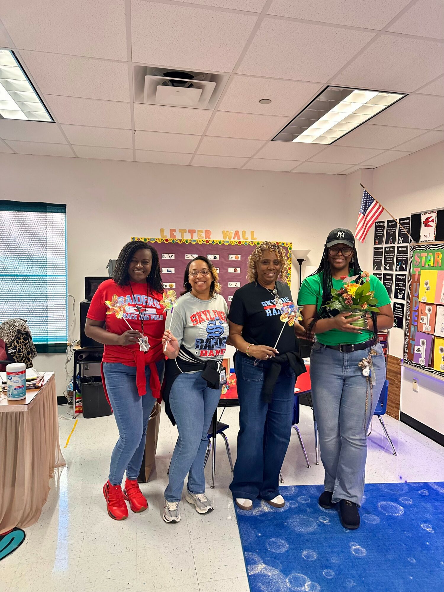 Four women standing in a classroom, holding books and smiling, with colorful posters and an American flag in the background.