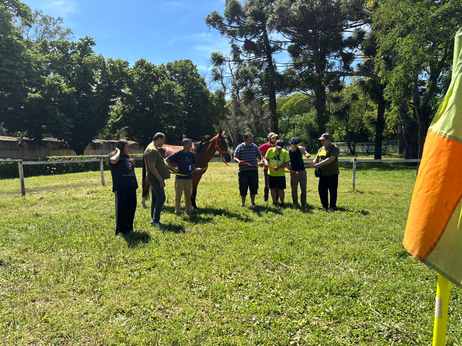 Group of people standing outdoors on grass, some holding papers, with trees and a flag partially visible on the right.