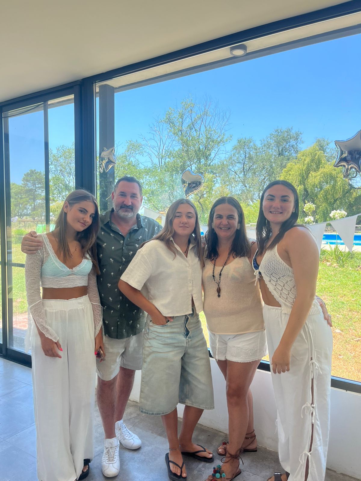 Five people standing indoors near large window, outdoor trees and blue sky visible, smiling and posing together.