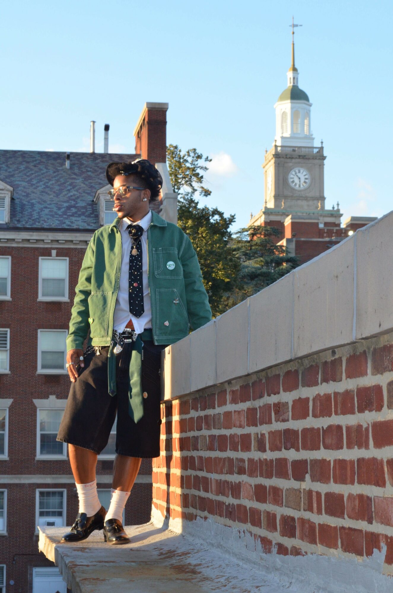 Young person with curly hair wearing glasses, green jacket, black shorts, and tie, standing on a brick wall outdoors.