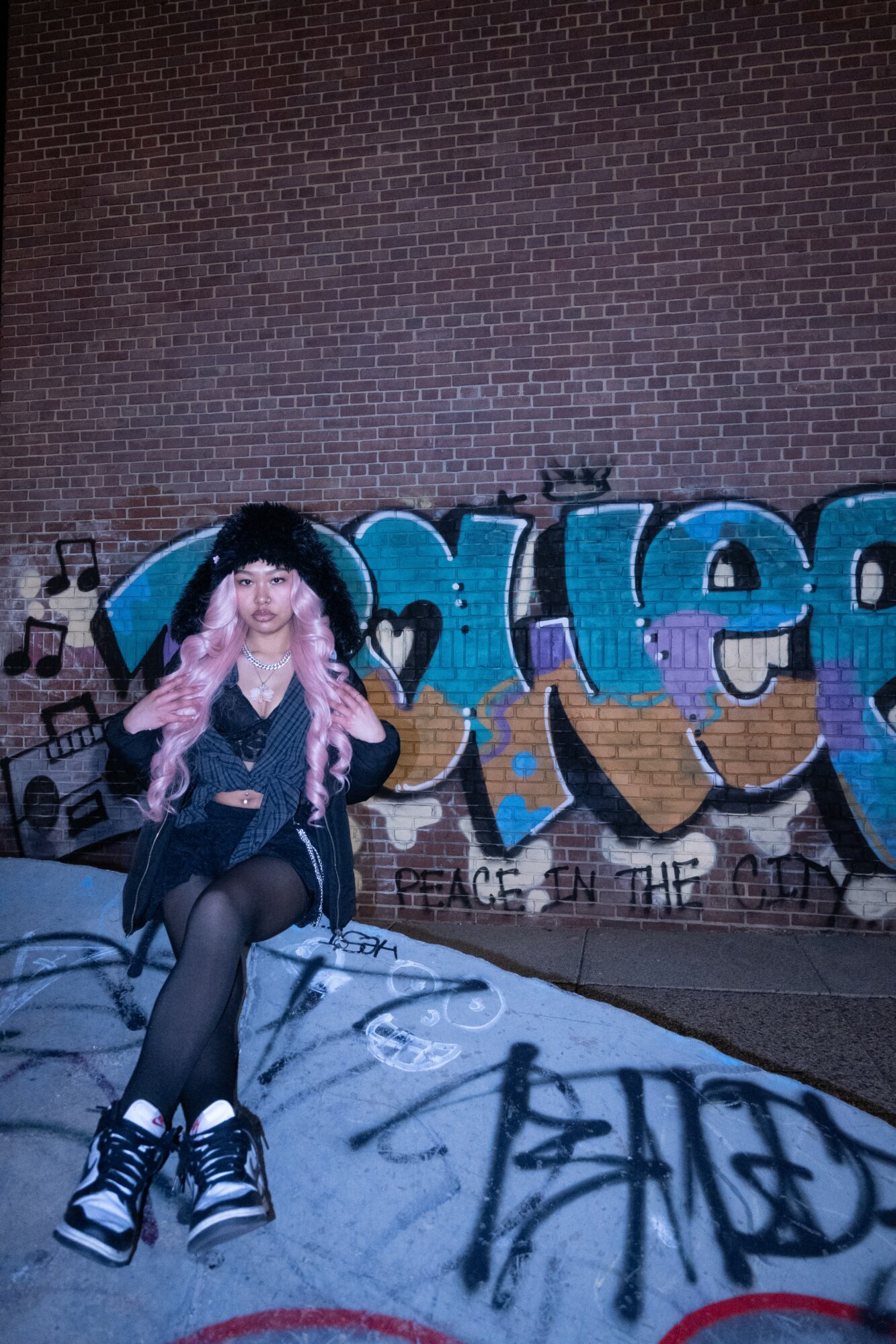 Young woman with pink hair sitting on the ground in front of a graffiti wall, wearing a black jacket and sneakers.