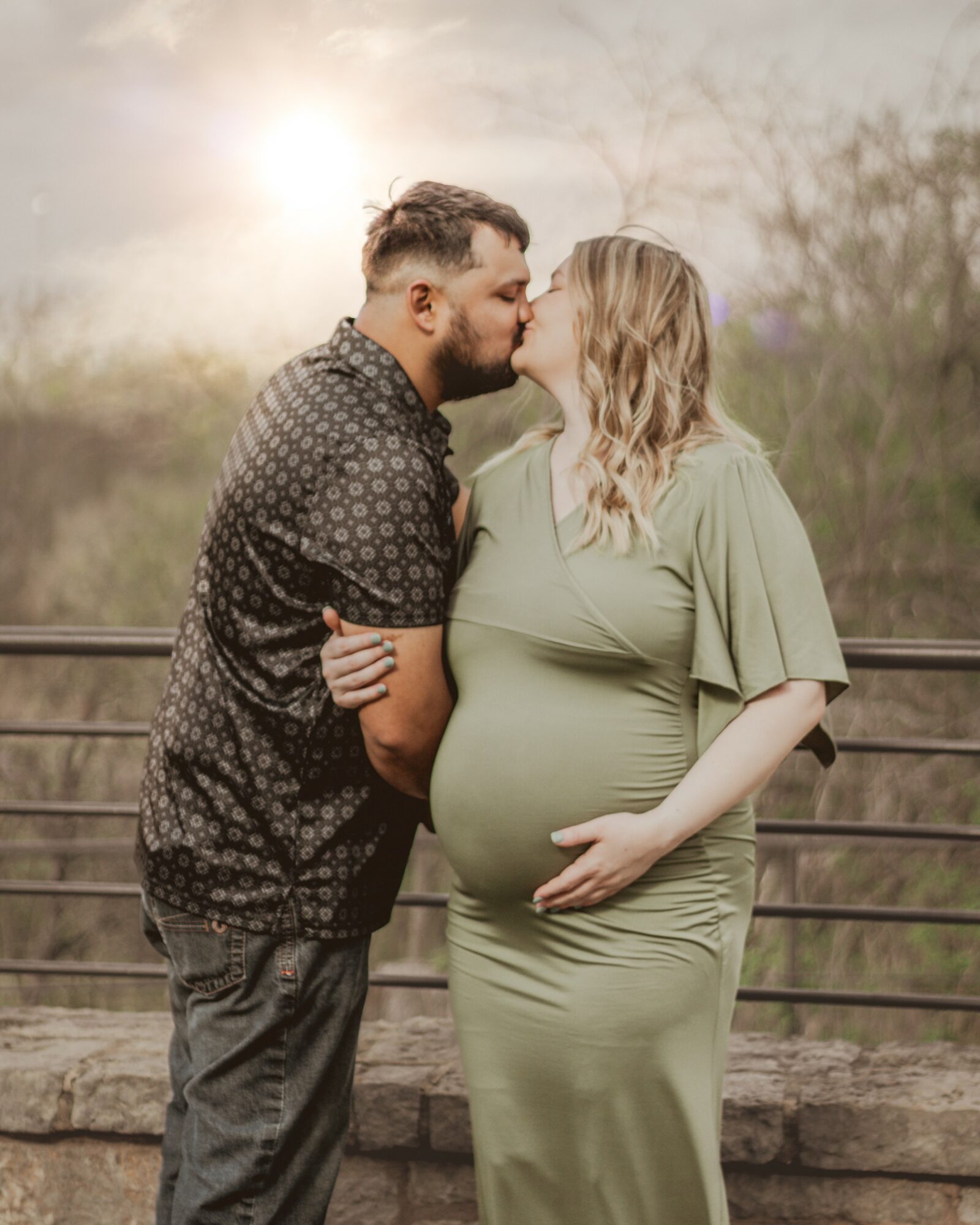 Man and pregnant woman standing close, touching foreheads outdoors with sunlight in background.