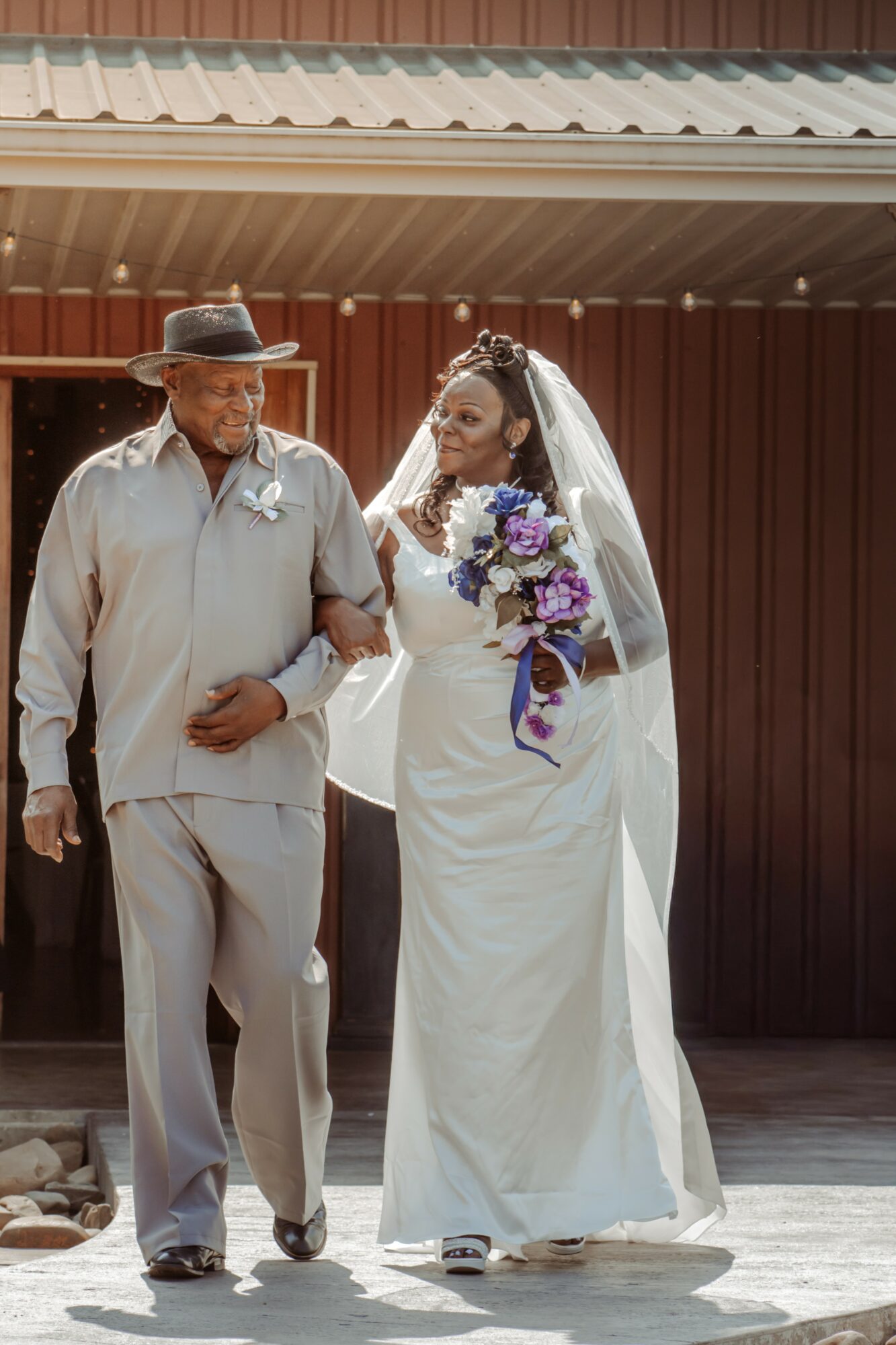 Bride in white wedding dress holding bouquet walking with an older man outside a building.