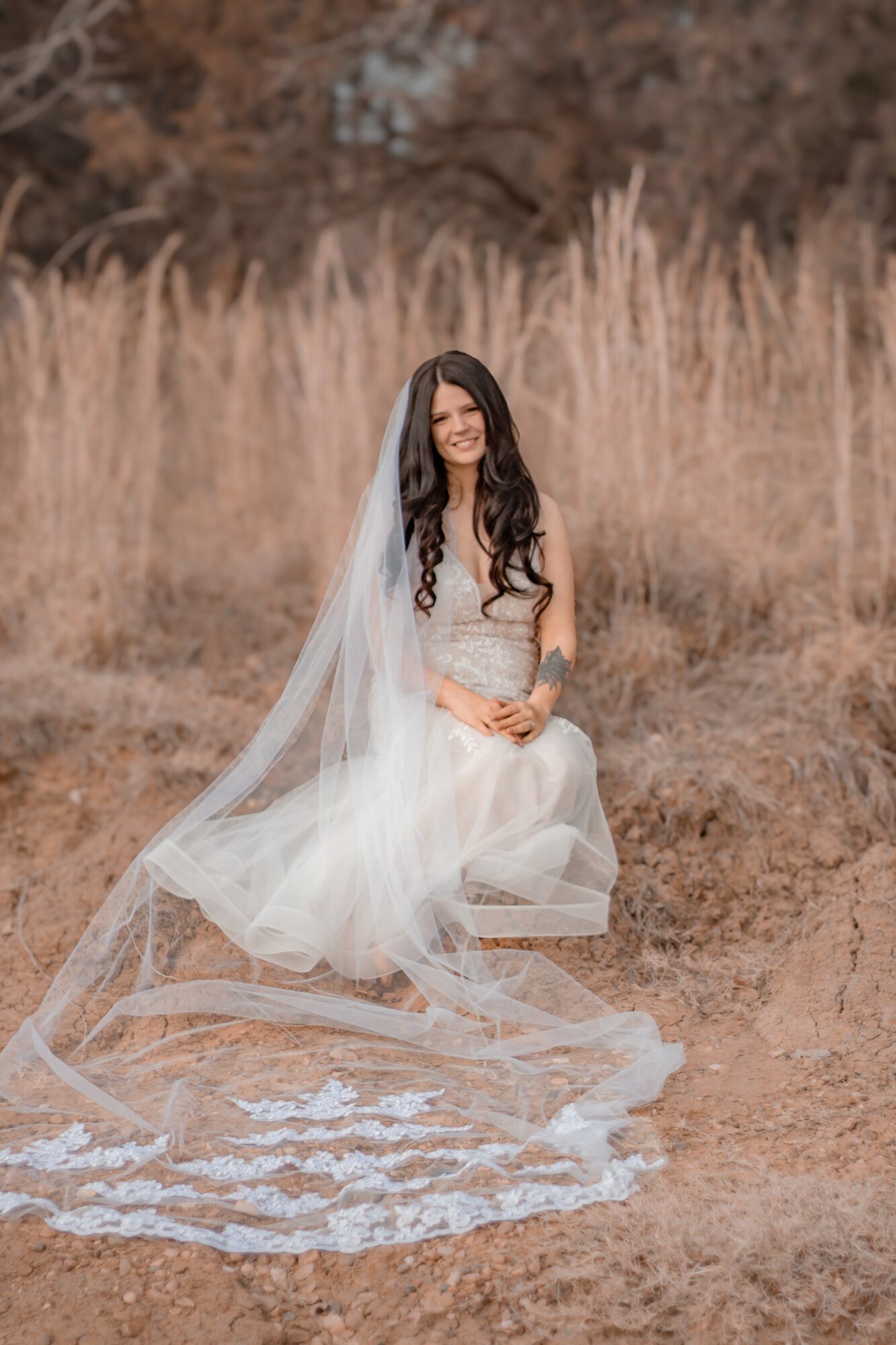 Woman in wedding dress sitting outdoors with tall grass background, veil flowing on ground.