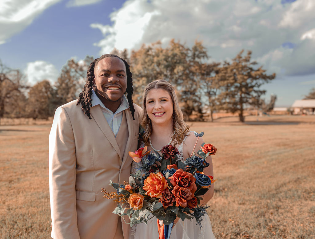 A man and woman stand outdoors holding a bouquet of flowers, smiling, with trees and a cloudy sky behind them.