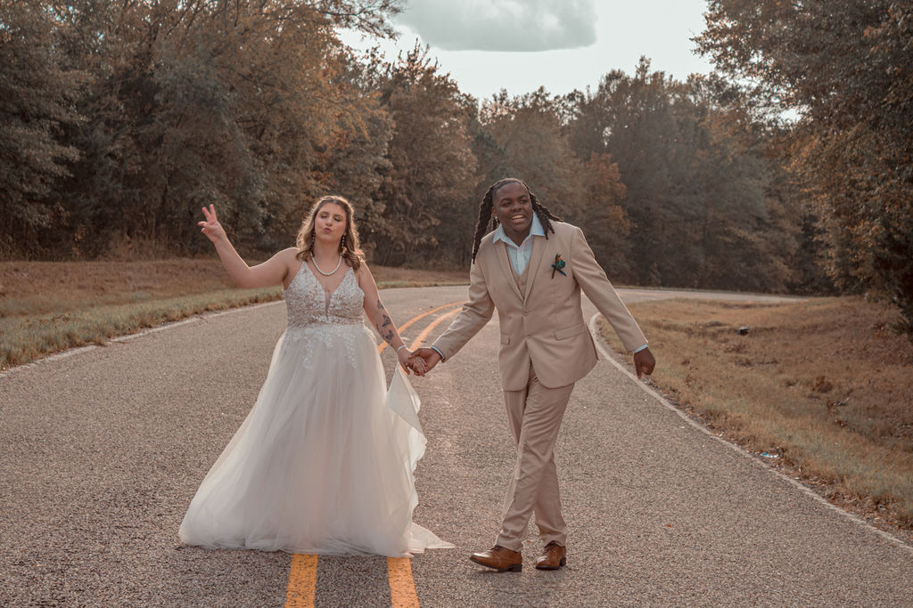 Couple holding hands walking on a road surrounded by trees, woman in a white dress and man in a beige suit.