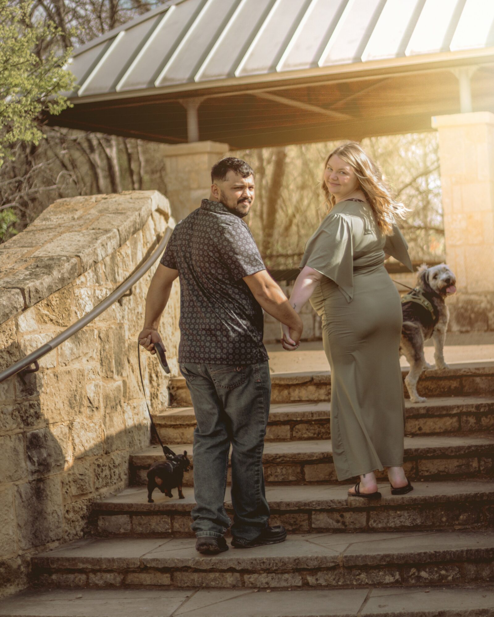 A couple holding hands on outdoor stairs with a dog nearby, in a park-like setting with trees and a stone wall.