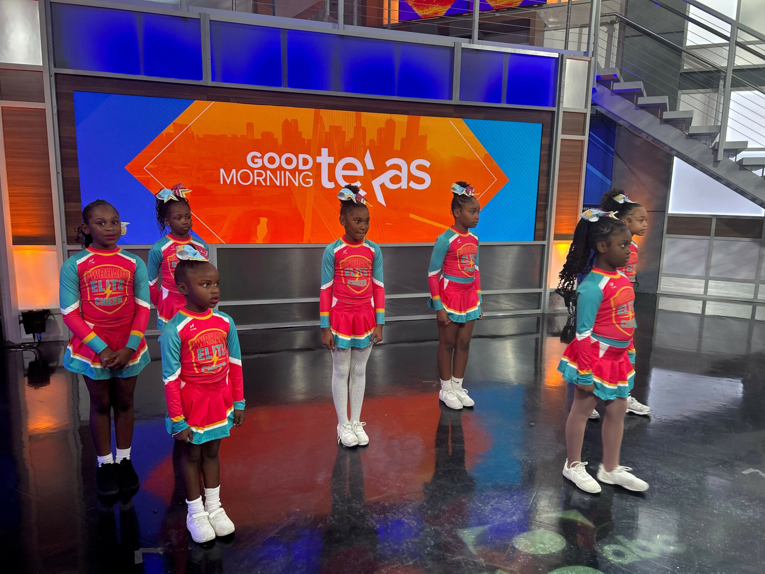 Six children stand in a row indoors, wearing colorful outfits, with a large screen behind them displaying 'Good Morning Texas'.