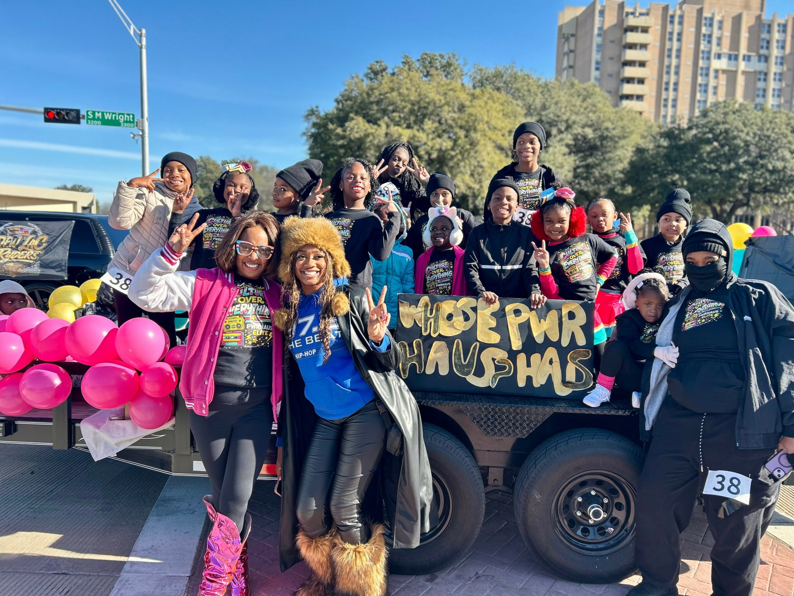 Group of people gathered around a truck holding pink balloons and a sign, outdoors with trees and buildings in background.