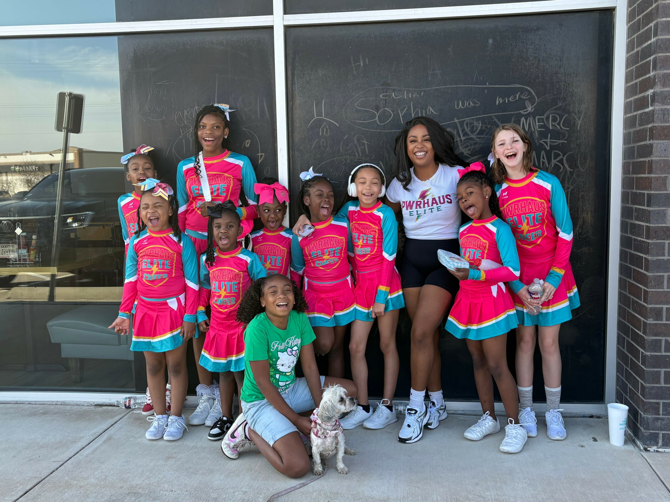 Group of young cheerleaders and two women standing outside near a blackboard, smiling, with a small dog.