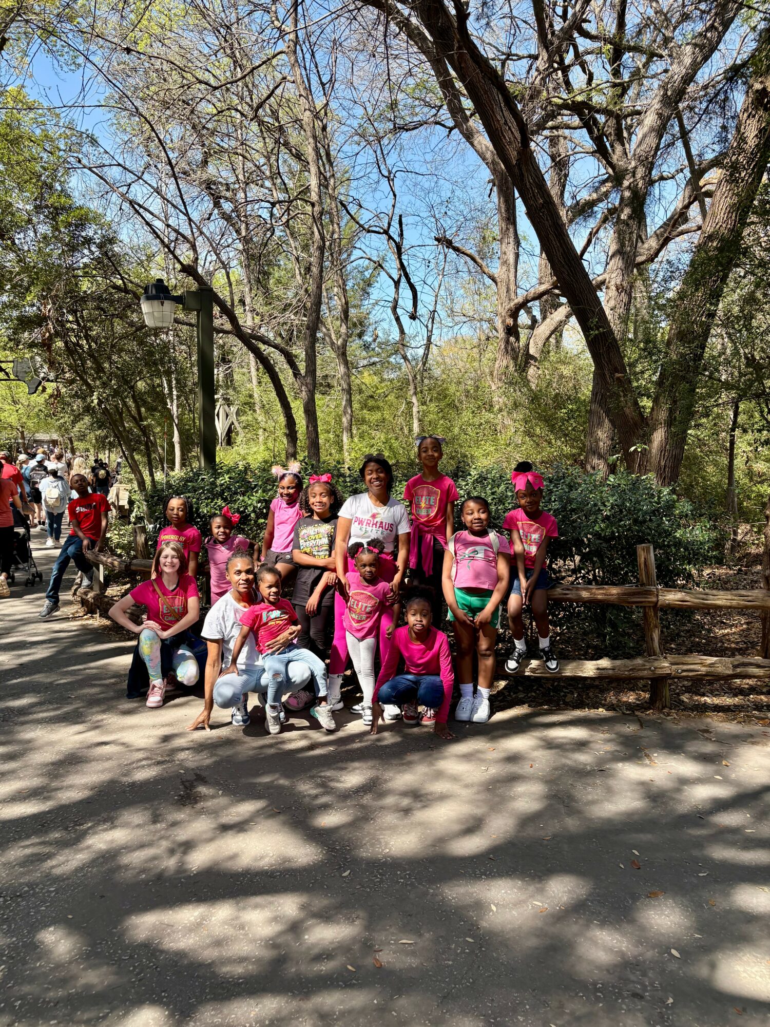 Group of children and adults in a park with trees and a pathway, some children wearing pink shirts and hats.
