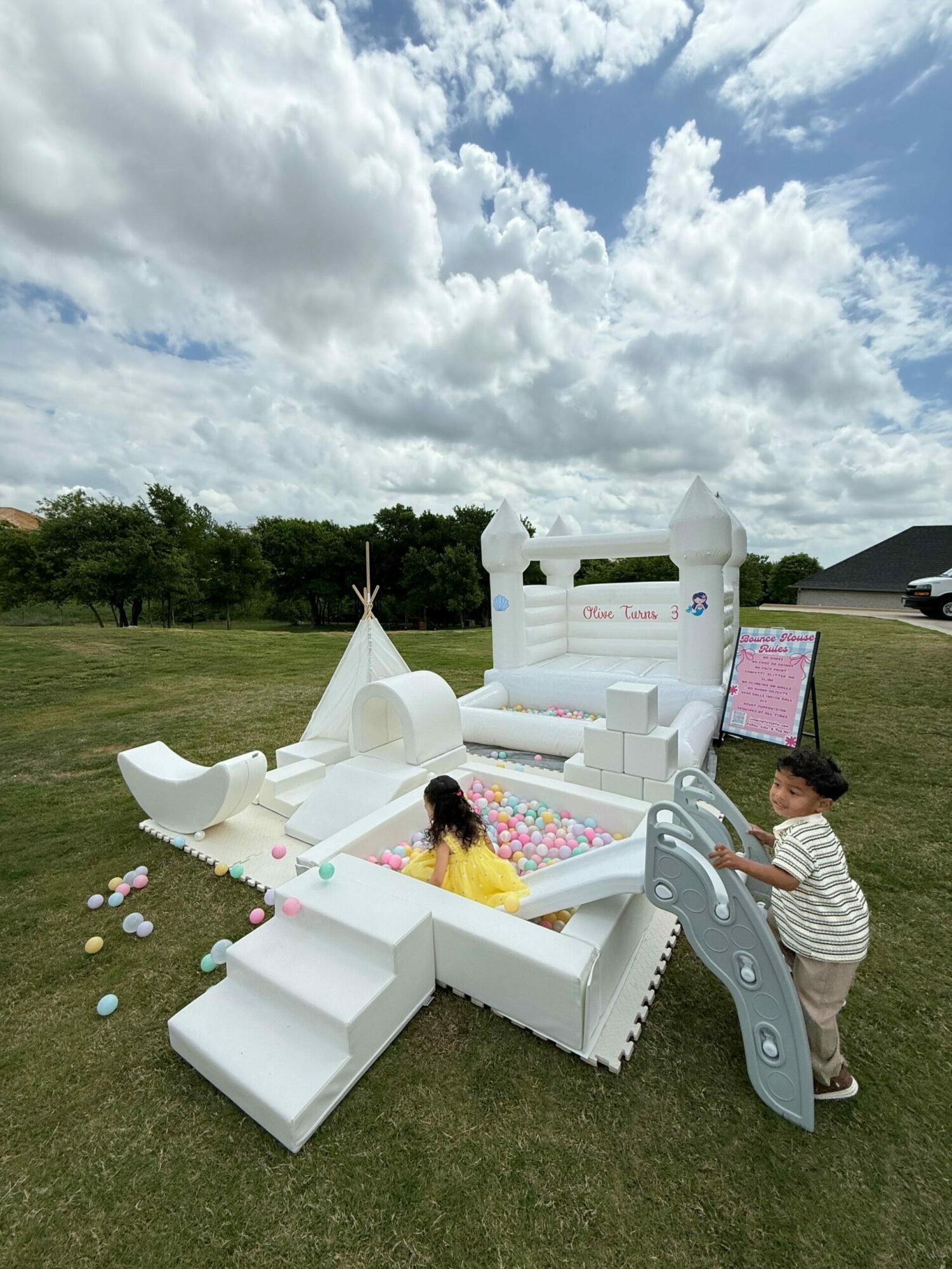 Child playing with a white and pink outdoor ice cream stand on grass, with clouds and trees in the background.