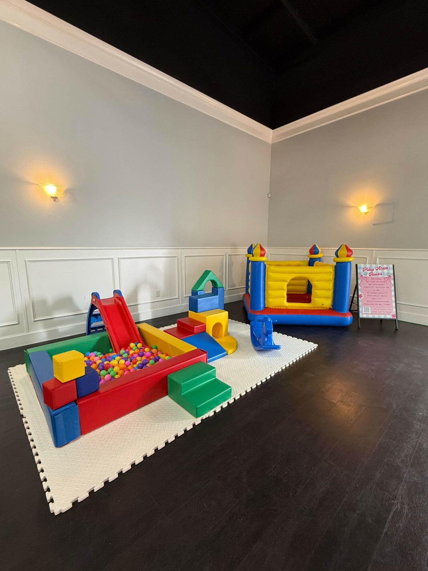Indoor play area with colorful inflatable bounce house, slide, and ball pit on white foam mat, dark floor, and light walls.