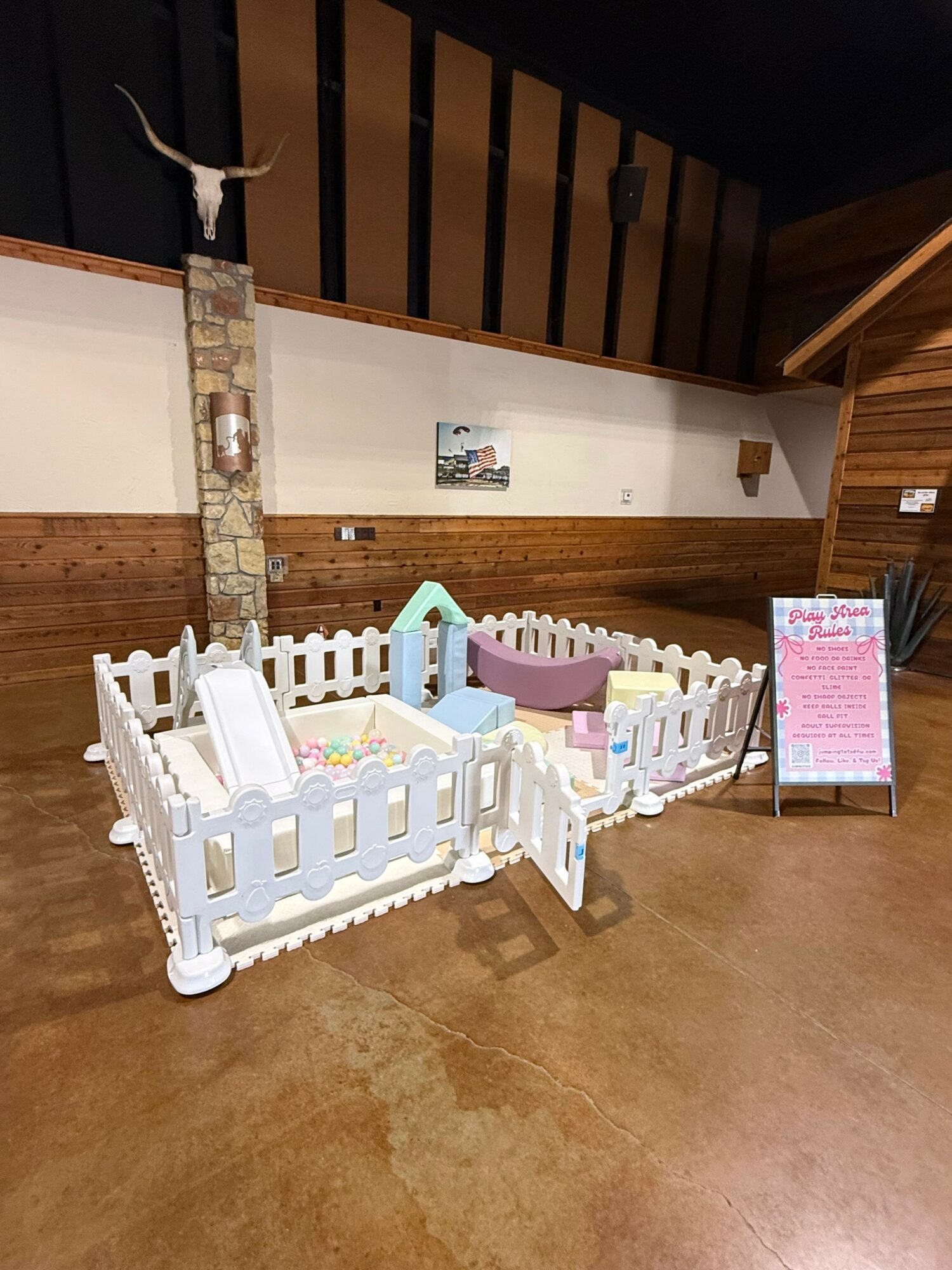 Indoor play area with white fence, colorful toys, and a pink chalkboard sign, against wooden walls and a stone pillar.