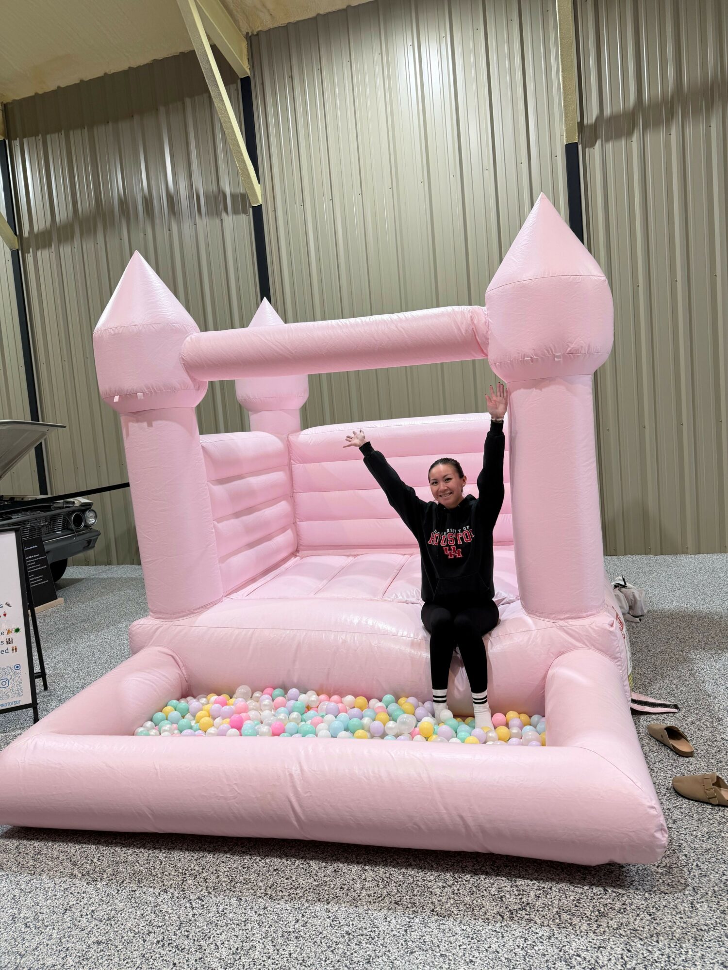 Child standing inside pink inflatable castle with ball pit, arms raised, in an indoor space with metal walls.