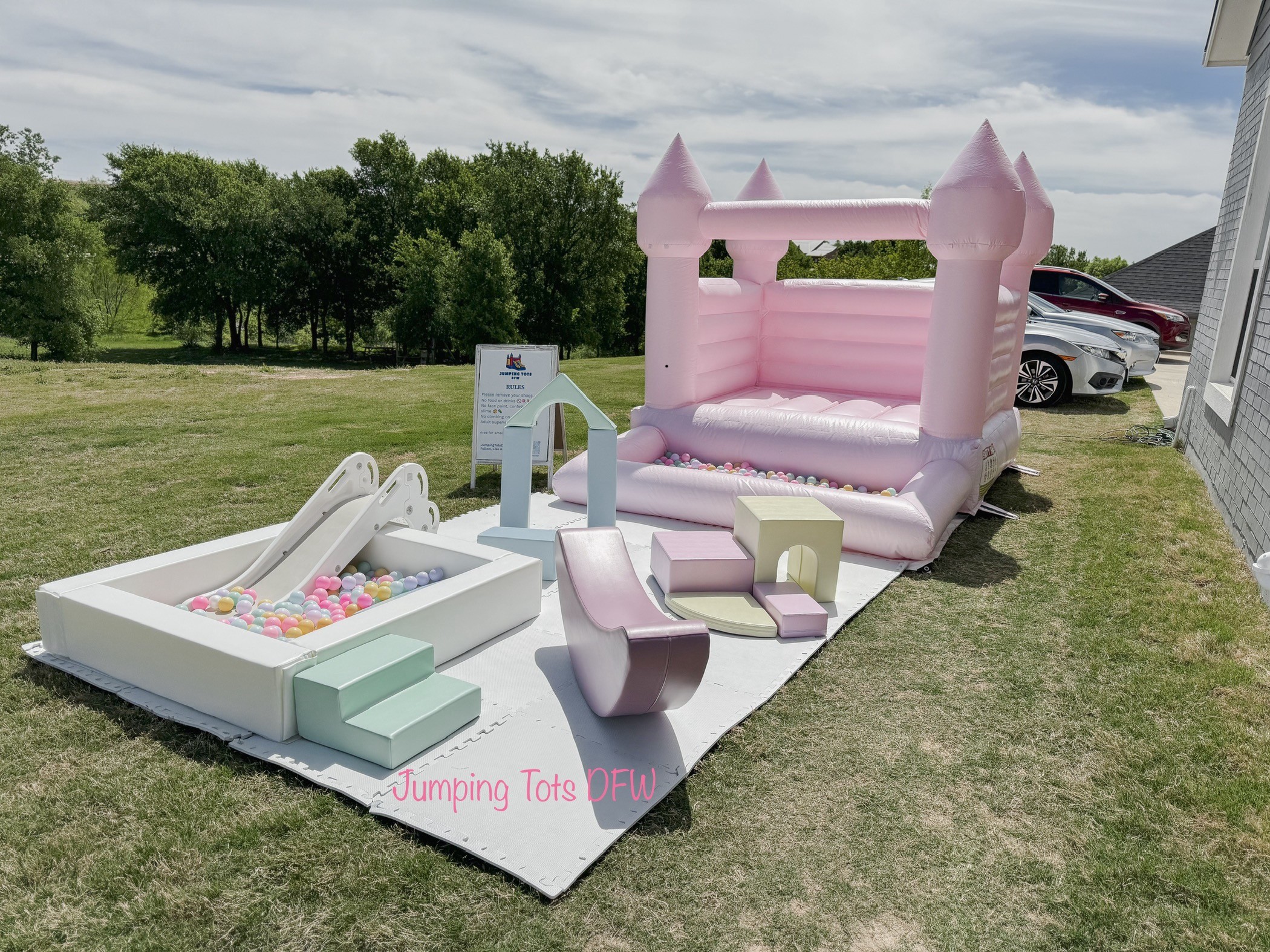 Pink inflatable castle with towers, a white inflatable slide, and a sandbox with toys on grass outdoors.