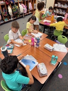 Children sit at tables in a classroom, working on assignments with books and supplies, with shelves of bags in background.