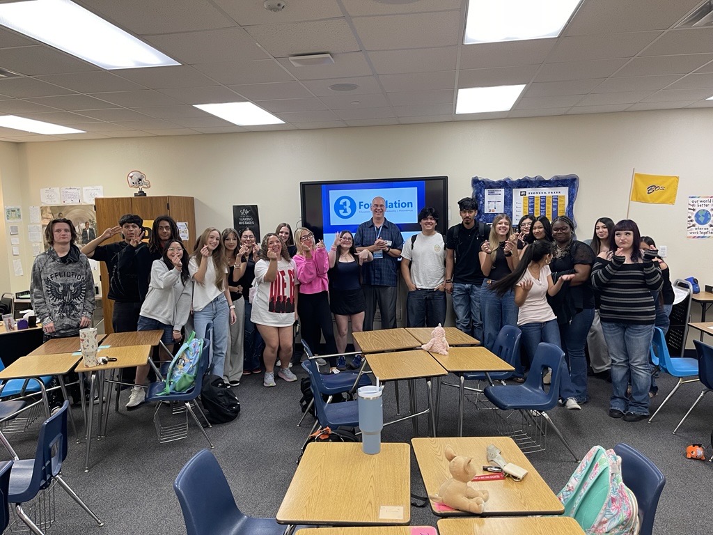 Group of people standing in a classroom, some waving, with desks and a large screen in the background.