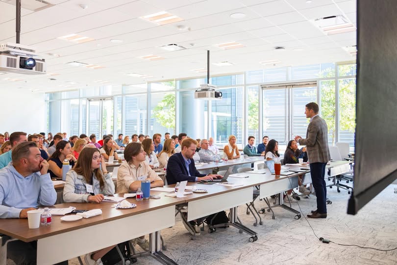A man presenting to a large group in a bright conference room with windows and ceiling lights.